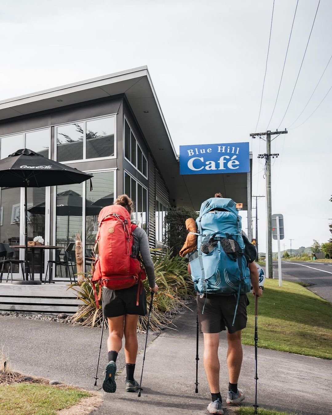 Two hikers with large backpacks approach the Blue Hill Café, a modern building with outdoor seating and a sign. The scene suggests a welcoming stop.