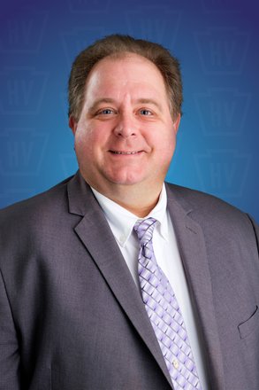 A professional man in a suit smiles against a blue background with a subtle pattern, conveying a welcoming and approachable atmosphere.
