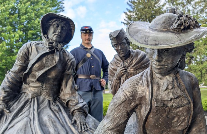 Bronze statues of three historical figures in period clothing, with a soldier in uniform standing behind them in a green park setting.