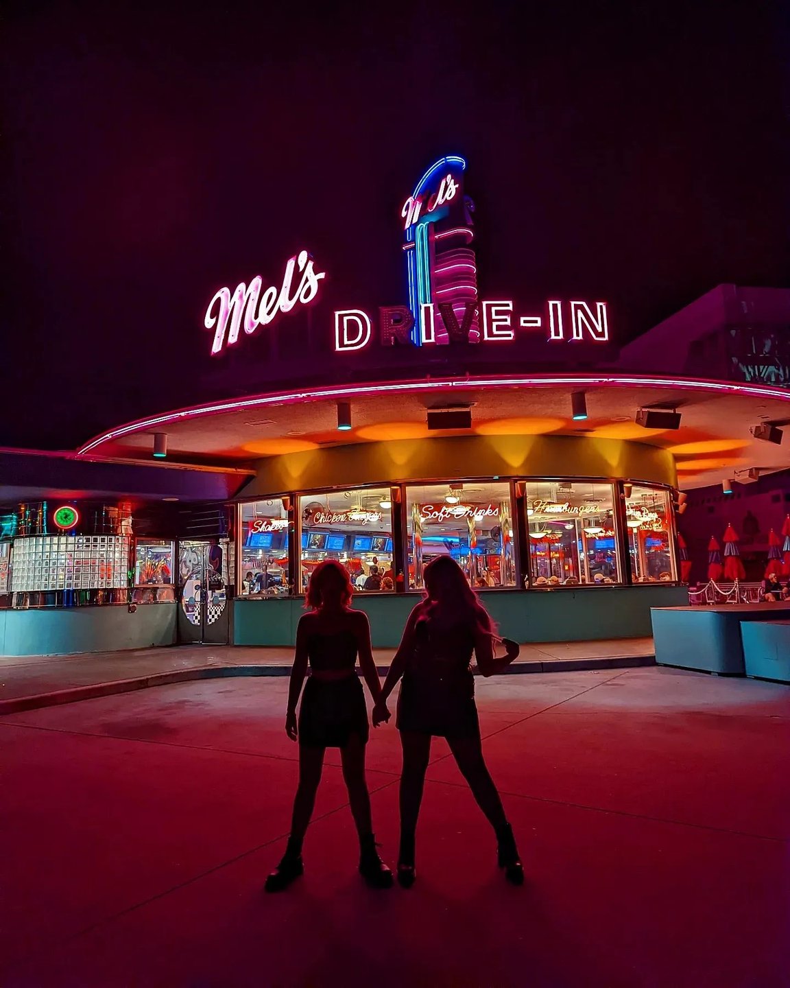 Silhouettes of two people holding hands in front of Mel's Drive-In, a vibrant diner with neon lights at night, creating a nostalgic atmosphere.