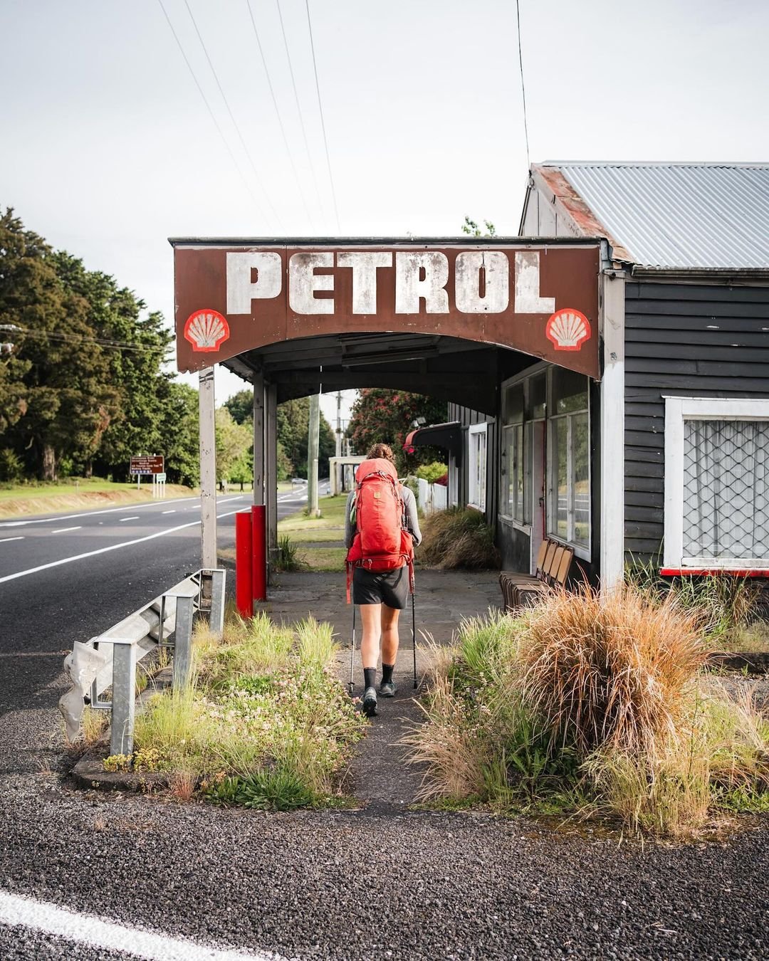 A hiker approaches an old petrol station with a weathered sign, surrounded by overgrown grass and trees along a quiet road.