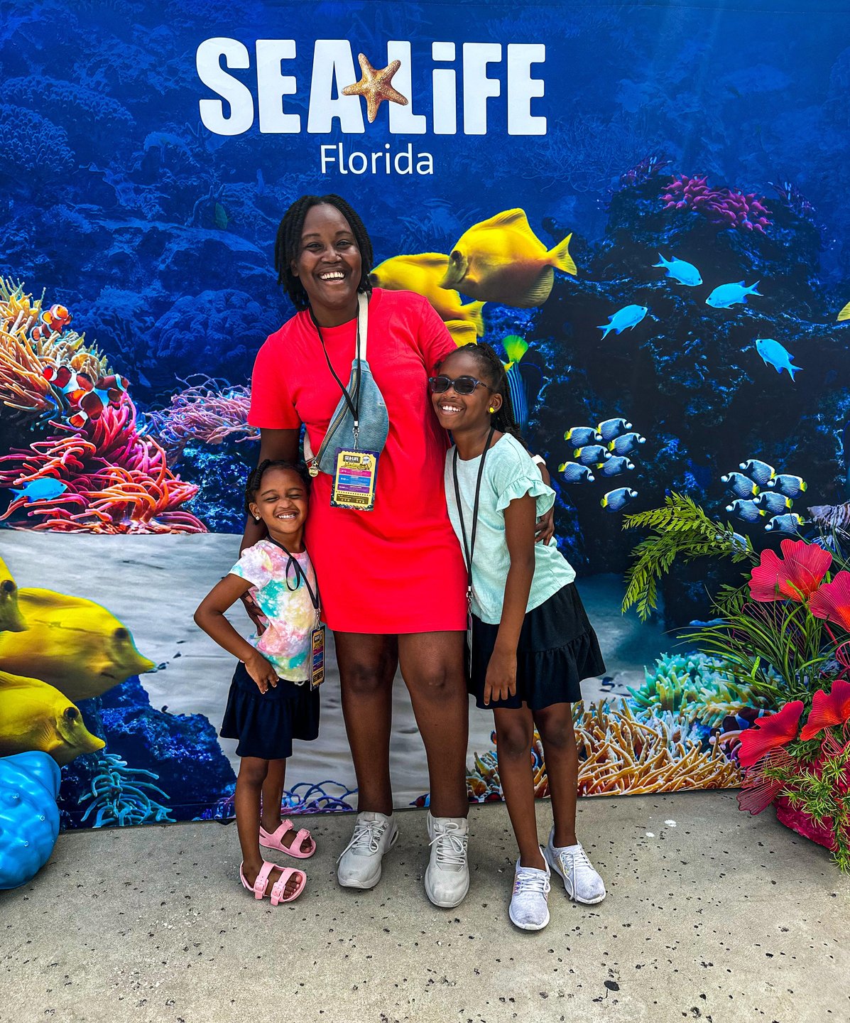 A woman and two girls pose in front of a vibrant underwater-themed backdrop featuring colorful coral and fish. The backdrop promotes SEA LIFE Florida.