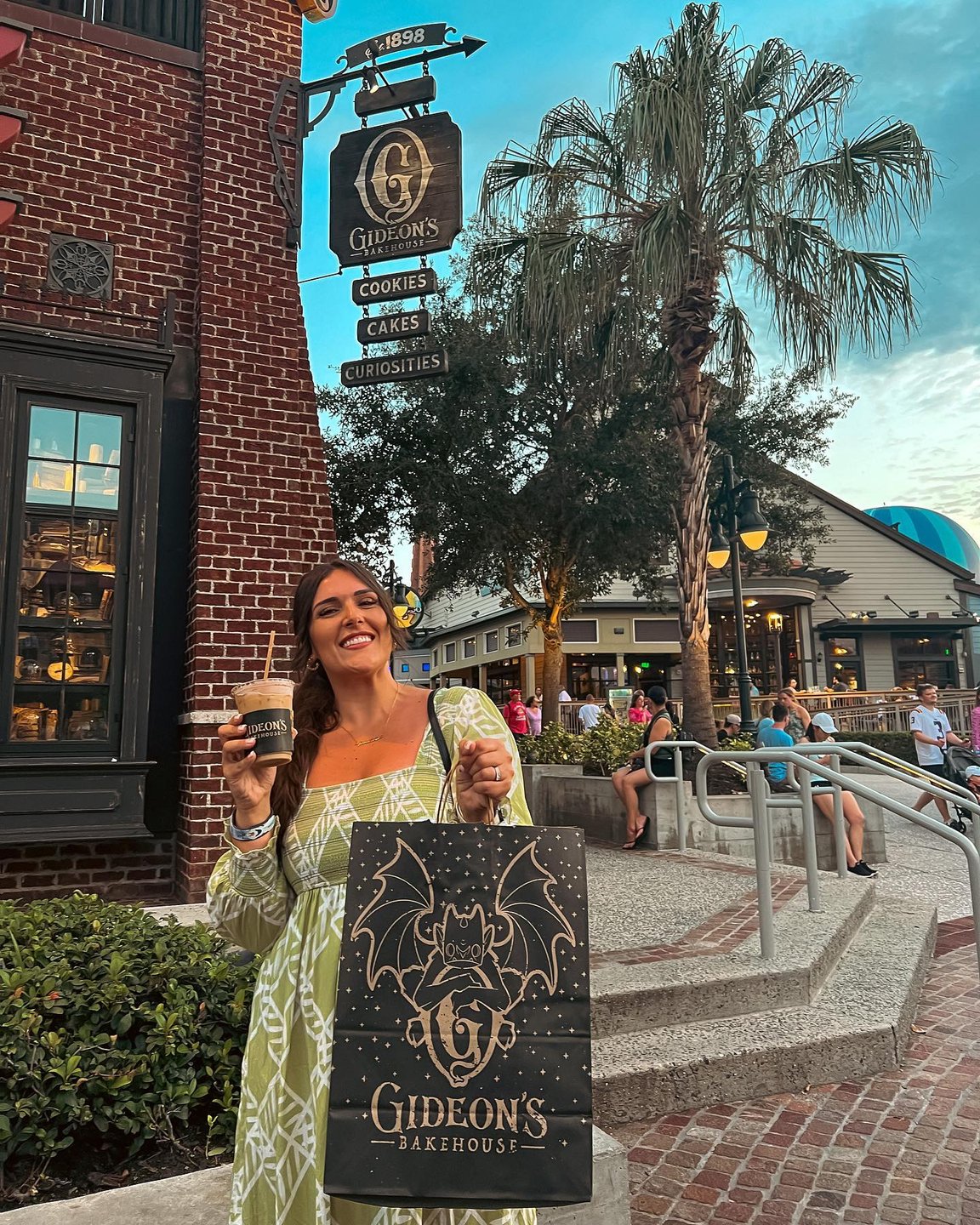 A woman holds a coffee and a shopping bag outside Gideon's Bakehouse, surrounded by palm trees and a lively street scene.
