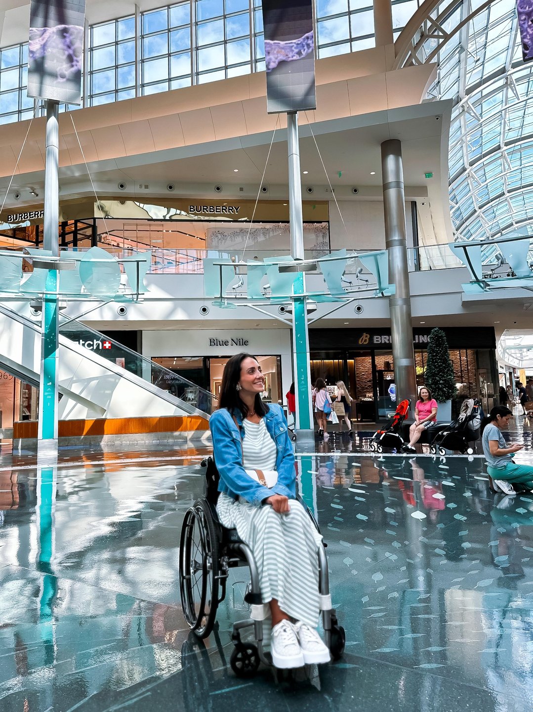 A woman in a wheelchair smiles in a spacious, modern shopping mall with large windows and various stores in the background.