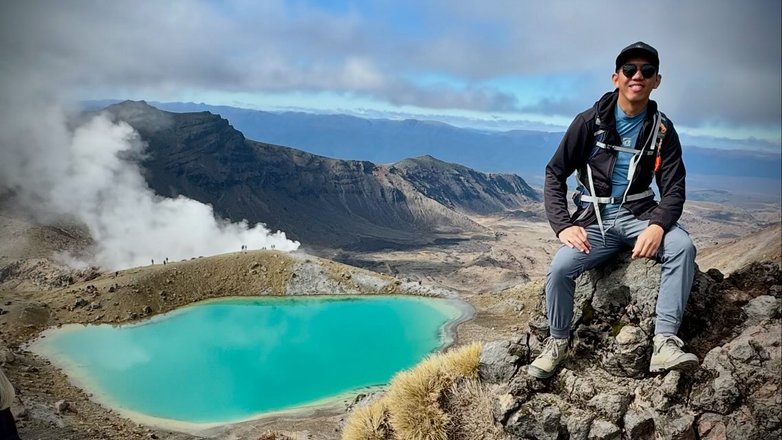 A hiker sits on a rocky outcrop overlooking a vibrant turquoise lake surrounded by rugged mountains and steam vents. The landscape is vast and dramatic.