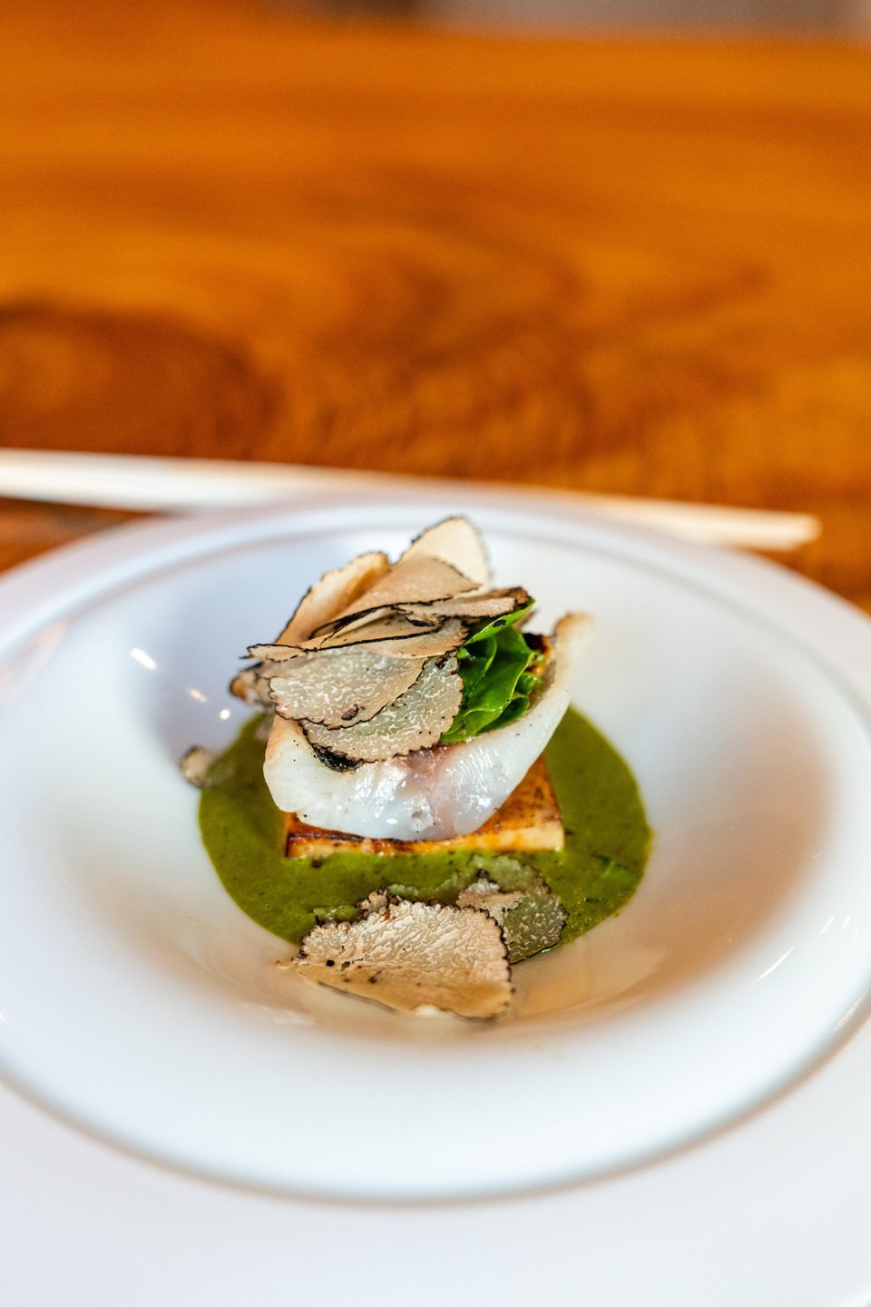 A gourmet dish featuring a delicate piece of fish atop a square of grilled bread, garnished with greens and truffle slices, served in a white bowl.