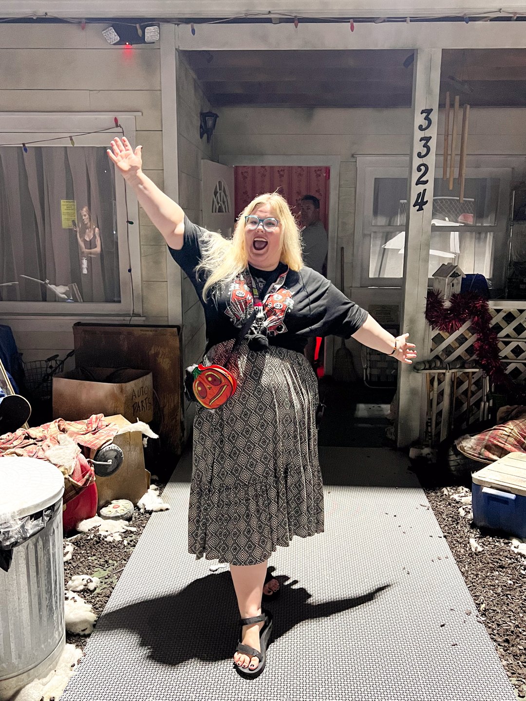 A person with blonde hair stands in front of a house, arms raised in excitement. The setting features a porch with various props and decorations.