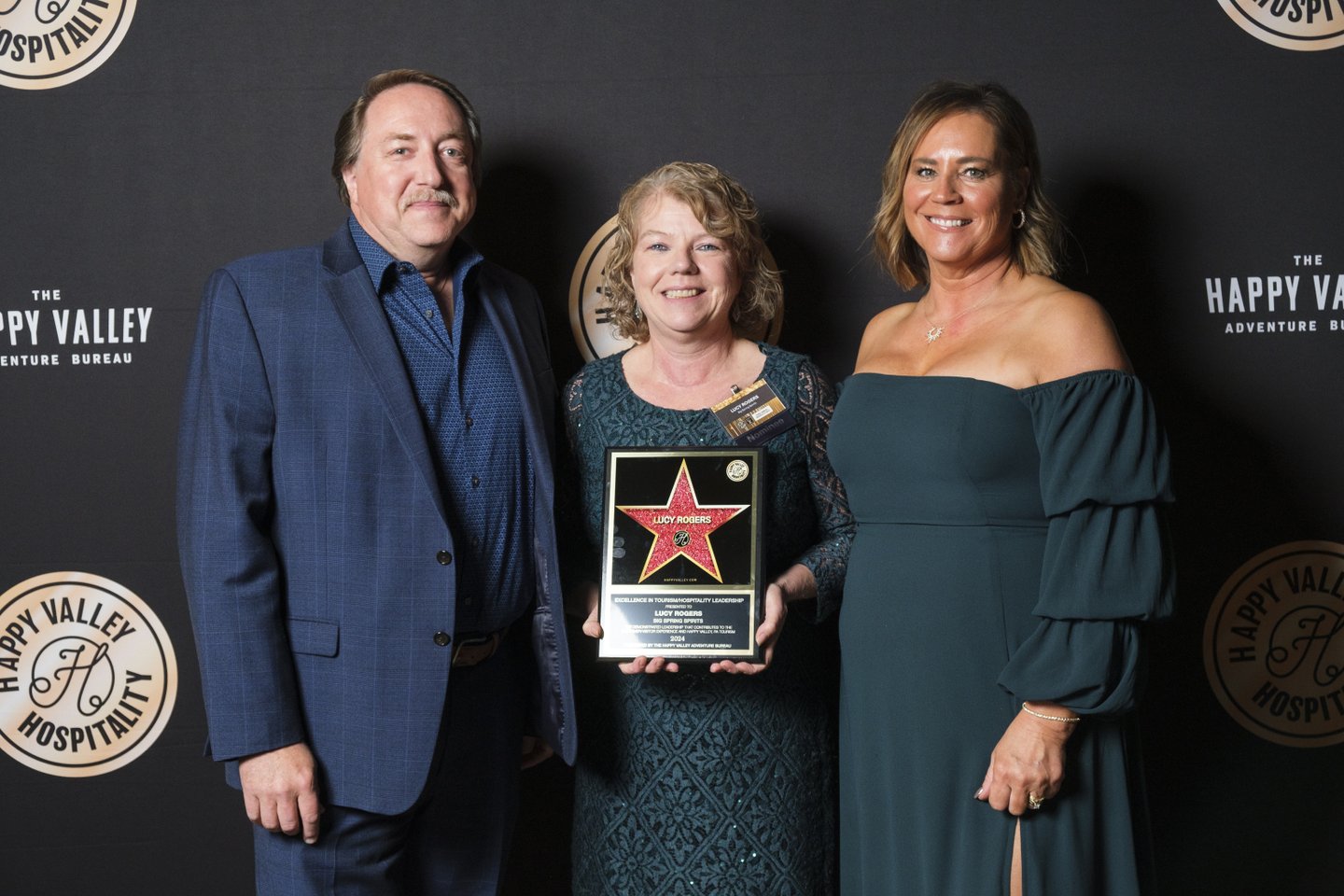 Three individuals stand together on a stage, holding an award plaque. The backdrop features logos for Happy Valley Hospitality.