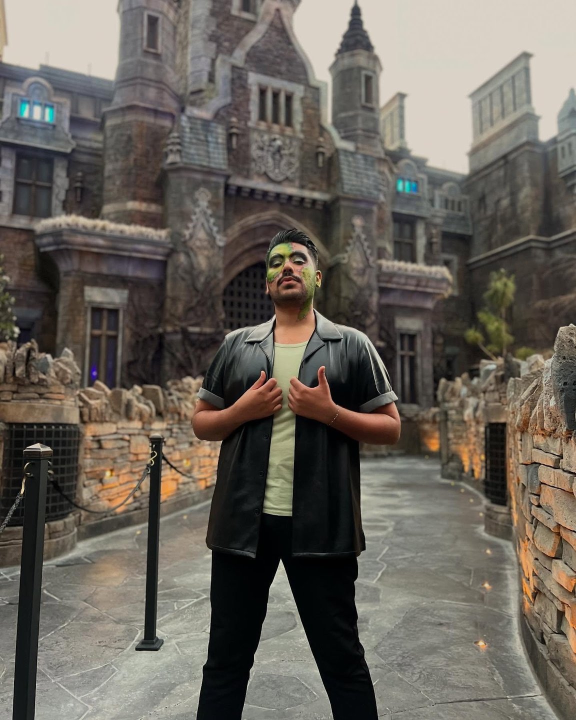 A person with green face paint stands confidently in front of a gothic-style building, surrounded by stone walls and dim lighting.
