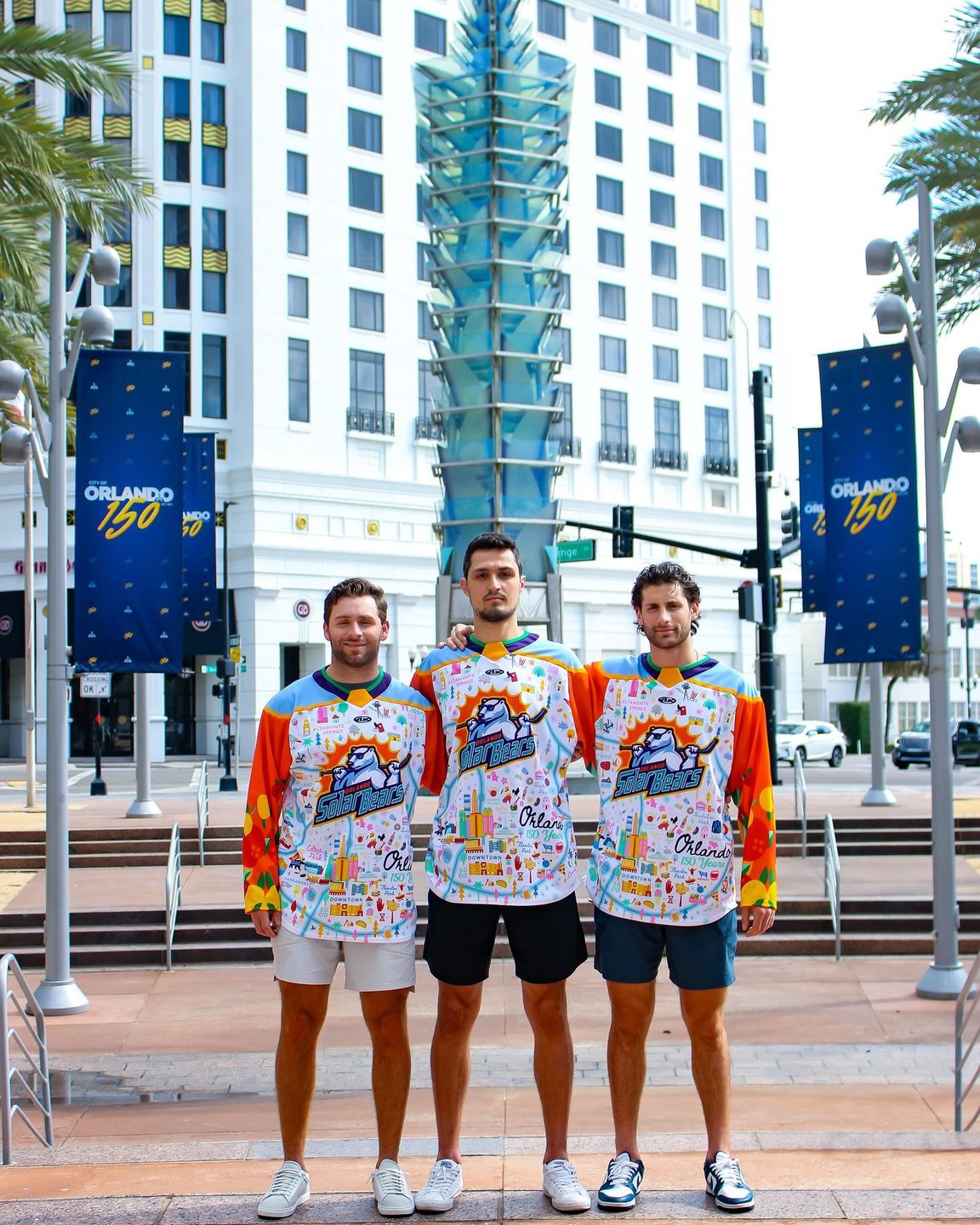 Three men stand in colorful, themed shirts in an urban setting, with a modern building and palm trees in the background. Banners promote "Orlando 150."