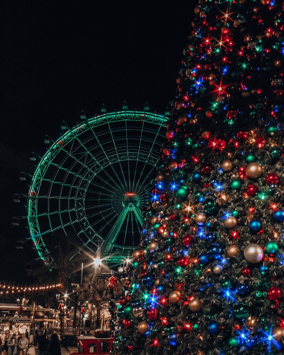 A brightly lit Christmas tree adorned with colorful ornaments stands in the foreground, with a large Ferris wheel illuminated in green behind it.
