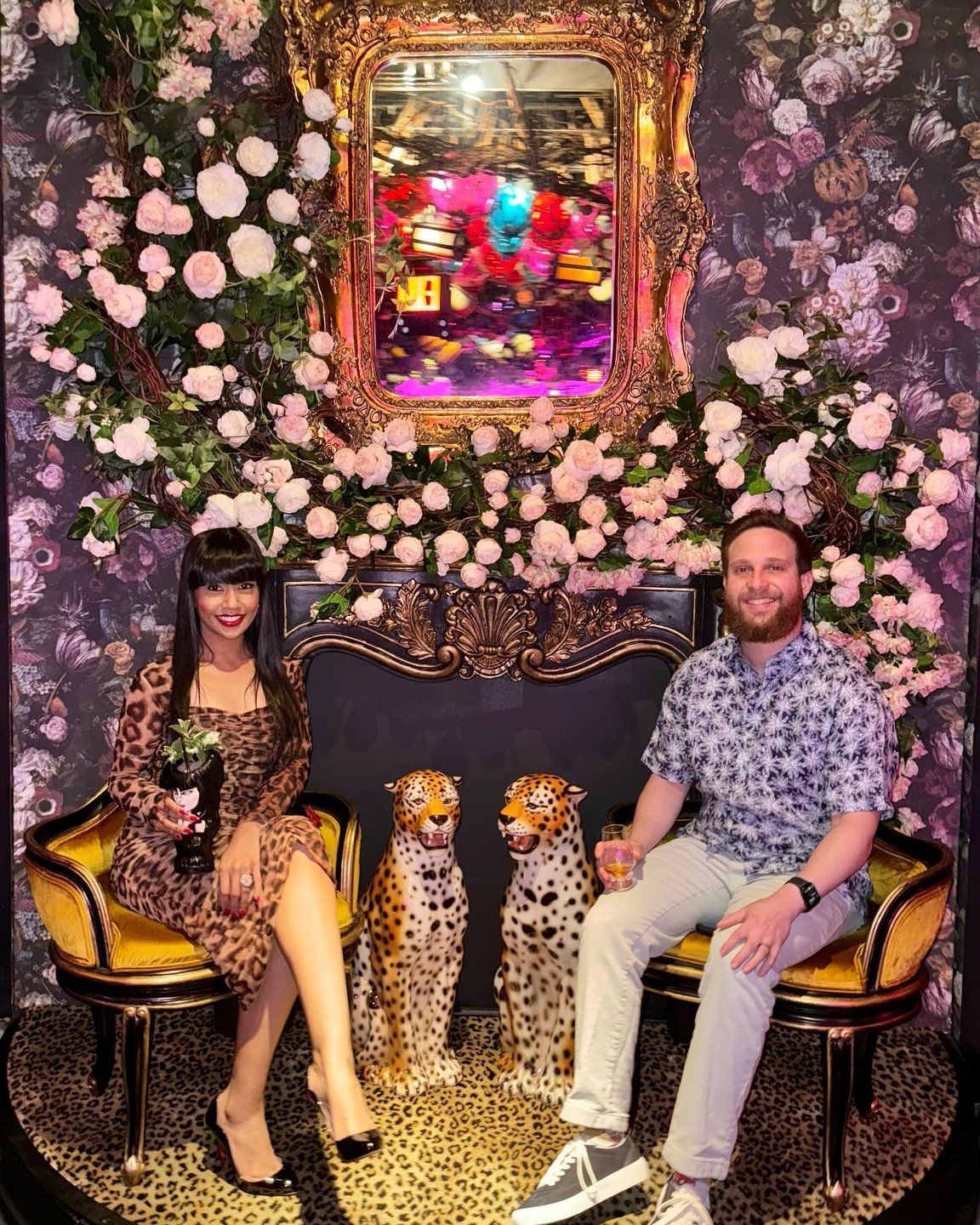 A stylish couple poses on ornate chairs surrounded by floral decor and leopard statues, set against a vibrant, patterned backdrop.