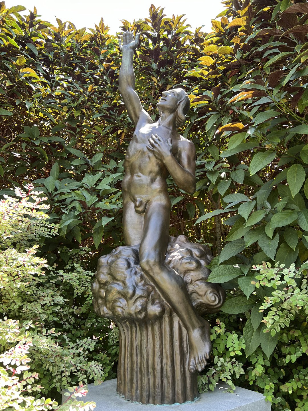 A bronze statue of a nude figure standing on a cloud, reaching upward amidst lush greenery and flowering plants. The scene evokes a serene garden atmosphere.