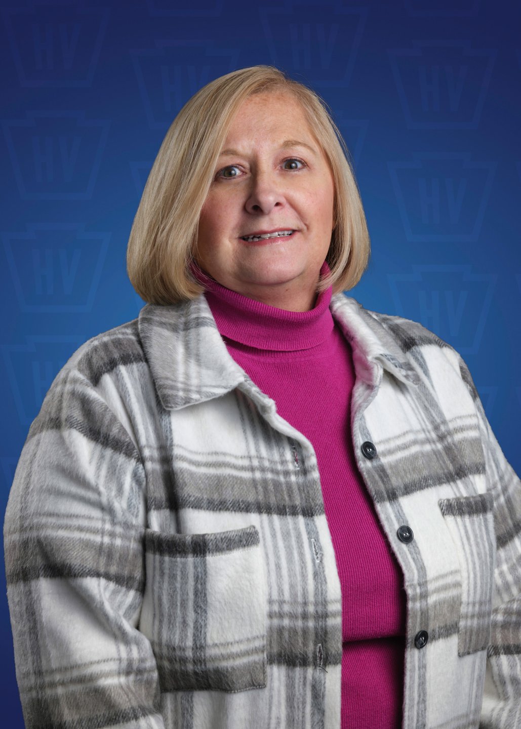 A woman with shoulder-length blonde hair smiles, wearing a plaid jacket over a pink top, set against a blue background.