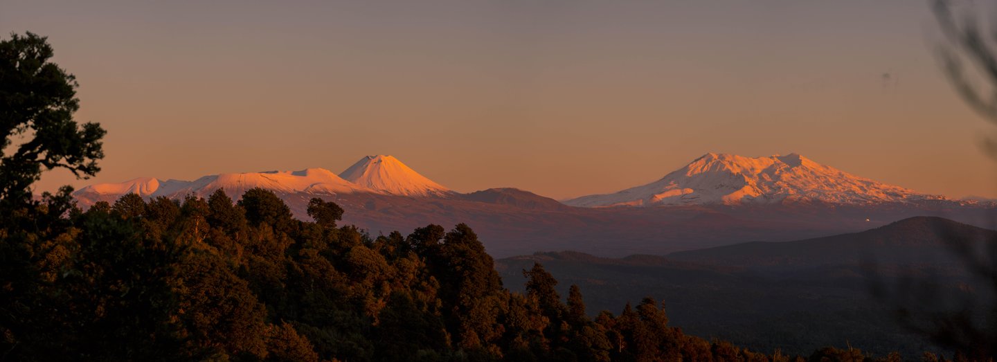 Snow-capped mountains rise majestically against a sunset sky, surrounded by lush forests and rolling hills, inviting exploration and adventure.