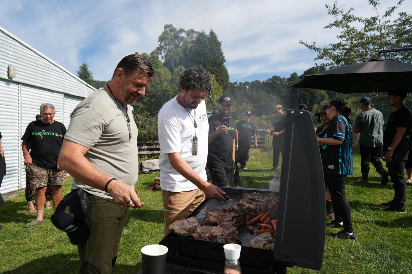 Two men grill meat and sausages outdoors at a gathering, surrounded by a diverse group of people enjoying the sunny day.