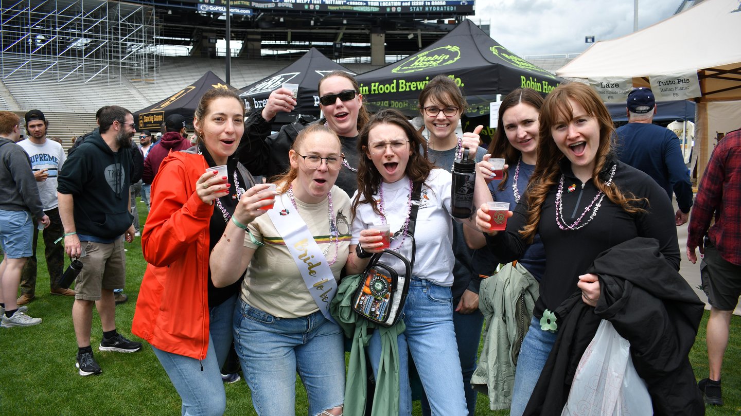A group of seven people, smiling and holding drinks, stands in a lively outdoor festival setting with tents in the background.