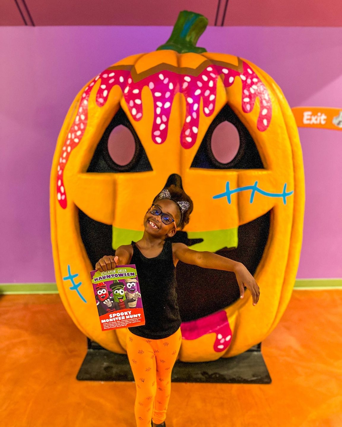 A child poses in front of a large, colorful pumpkin decoration with a playful expression, holding a festive sign. The vibrant background enhances the fun atmosphere.