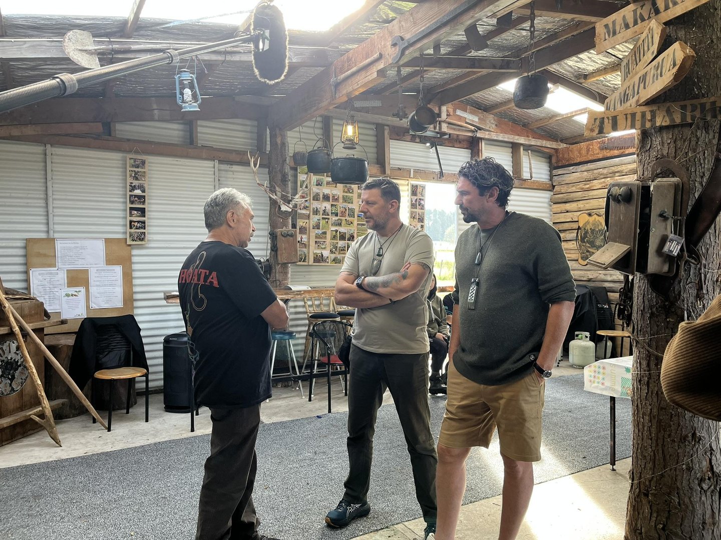 Three men engage in conversation inside a rustic, wooden interior with various photographs and artifacts on the walls. Tables and chairs are visible.
