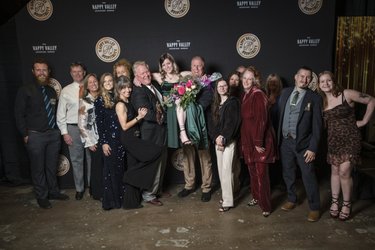 Group of people posing together in formal attire, celebrating an event. A woman in a green dress holds a bouquet. Happy Valley backdrop.