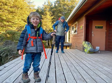 A young boy smiles on a wooden deck, holding a hiking pole. An adult prepares nearby, with backpacks and a cabin in the background.