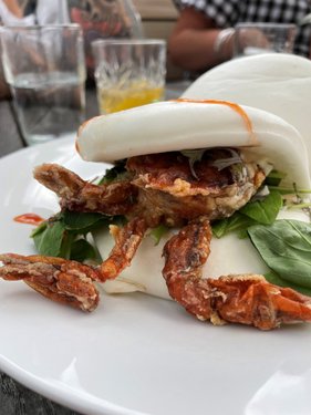 A close-up of a soft bao bun filled with crispy fried crab, garnished with fresh greens. Glasses of drinks are blurred in the background.