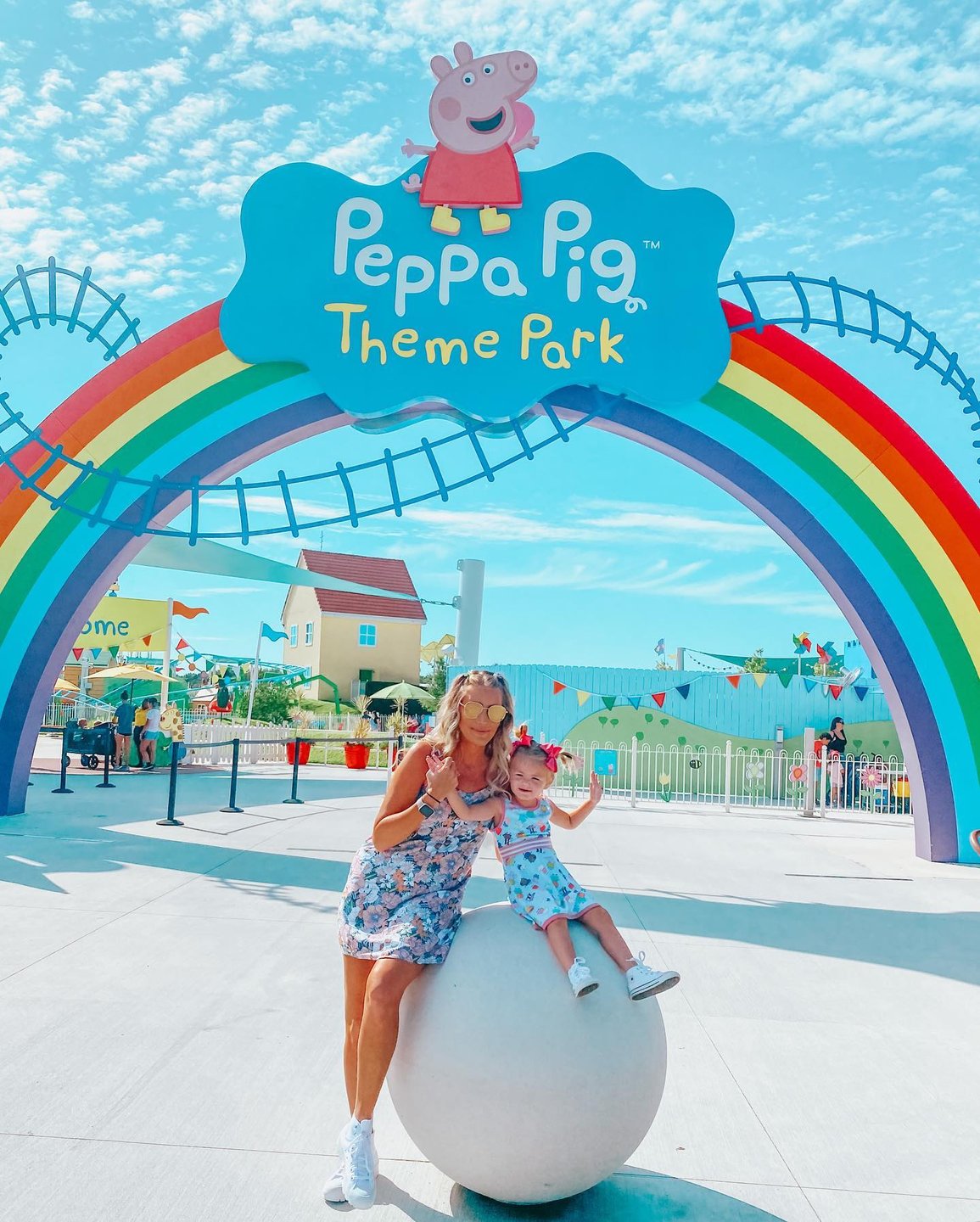 A colorful archway with "Peppa Pig Theme Park" signage welcomes visitors. A woman and child pose on a large white sphere, smiling.