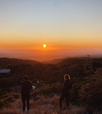 Two individuals stand on a hillside, silhouetted against a vibrant sunset over rolling mountains. The scene captures a serene landscape with lush greenery.