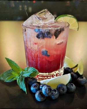 A refreshing cocktail with blueberries and lime, served over ice in a glass, garnished with mint leaves and lime wedges.