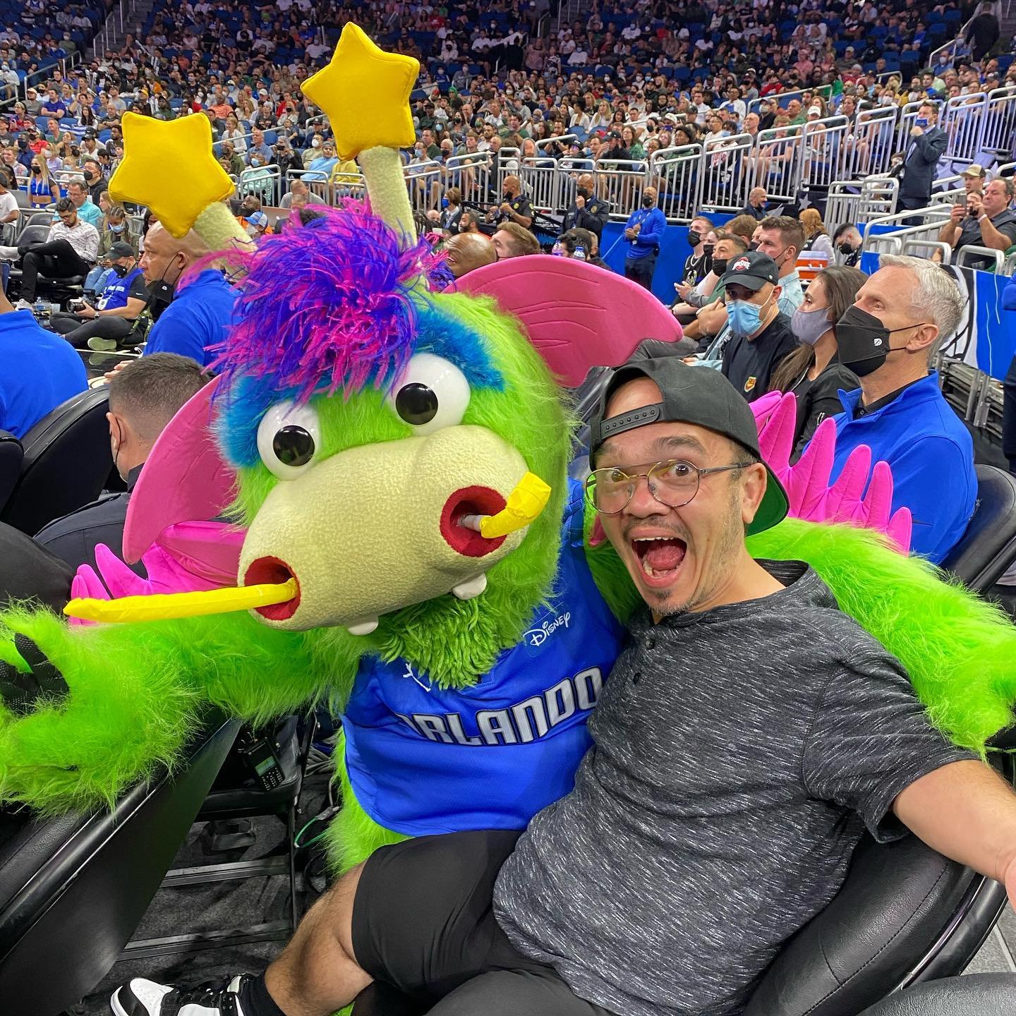 A vibrant green mascot with colorful features poses playfully with a smiling fan in a crowded sports arena, showcasing excitement and energy.
