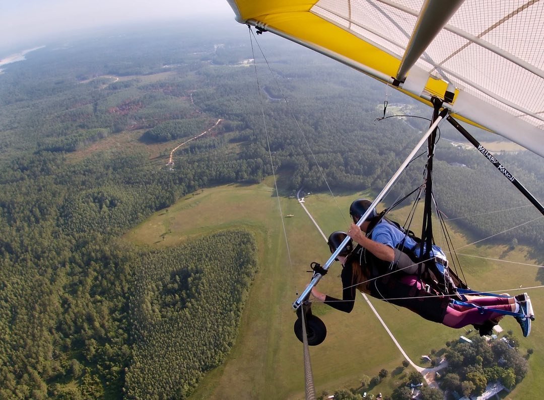 A person hang gliding over a lush green landscape, showcasing rolling hills and a winding path below. The scene captures the thrill of flight.