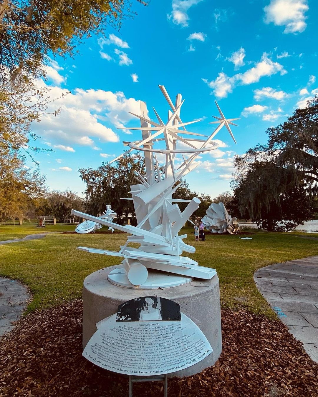 A white abstract sculpture stands in a green park, surrounded by trees and blue skies. Informational plaque at the base provides context.