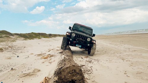 A black off-road vehicle navigates a sandy beach, positioned on a large log with ocean waves in the background under a cloudy sky.
