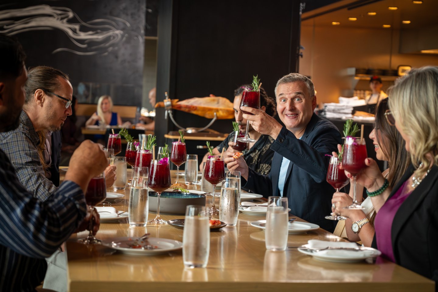 A lively dining scene featuring a group of people toasting with vibrant drinks at a modern restaurant. Plates and glasses are elegantly arranged on a wooden table.