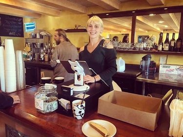 A barista stands behind a wooden counter in a cozy café, smiling while operating a register. Coffee cups and a slice of cake are visible.