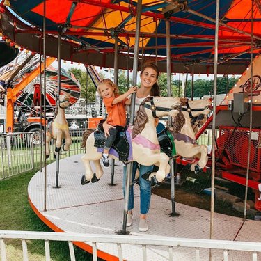 A carousel with colorful horses, featuring a woman and a child enjoying the ride. Brightly colored tents and attractions in the background.