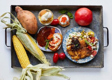 A metal tray holds a variety of dishes: fried fish, corn, sweet potato, and fresh vegetables, showcasing vibrant local cuisine.