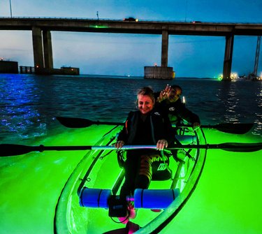 Two people kayak in glowing green water under a bridge at dusk, with city lights reflecting in the background. The scene captures an adventurous evening activity.