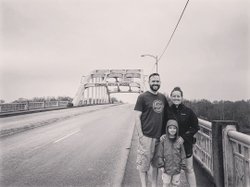 A family stands on a bridge with "Edmond Pettus Bridge" signage. The road is empty, surrounded by trees under a cloudy sky.