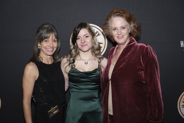 Three women pose together in front of a dark backdrop, smiling. They wear elegant outfits, suggesting a formal event or celebration.