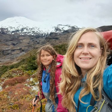 A woman and a girl smile for a selfie while hiking in a mountainous landscape with snow-capped peaks and colorful vegetation.