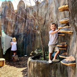 Two children play in a natural-themed outdoor space. One climbs a log structure while the other interacts with a musical installation.