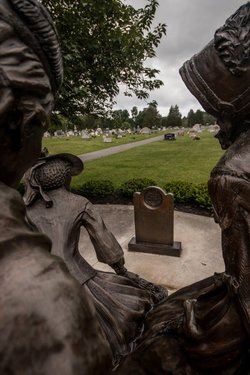 Bronze sculptures of two women seated, gazing at a gravestone in a serene cemetery. Lush greenery and gravestones are visible in the background.