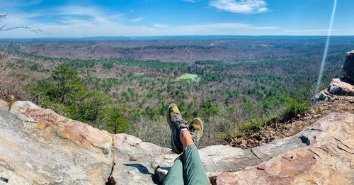 A person sits on a rocky ledge, legs crossed, overlooking a vast green valley and distant hills under a clear blue sky.