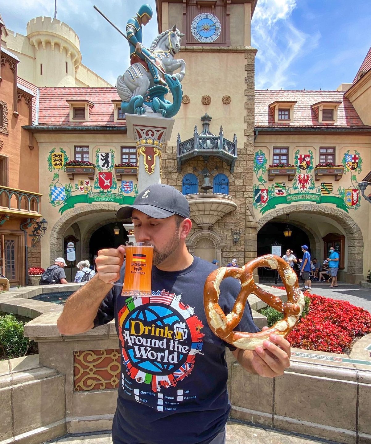 A man holds a large pretzel and a drink in front of a colorful, medieval-style building with flags and floral decorations.