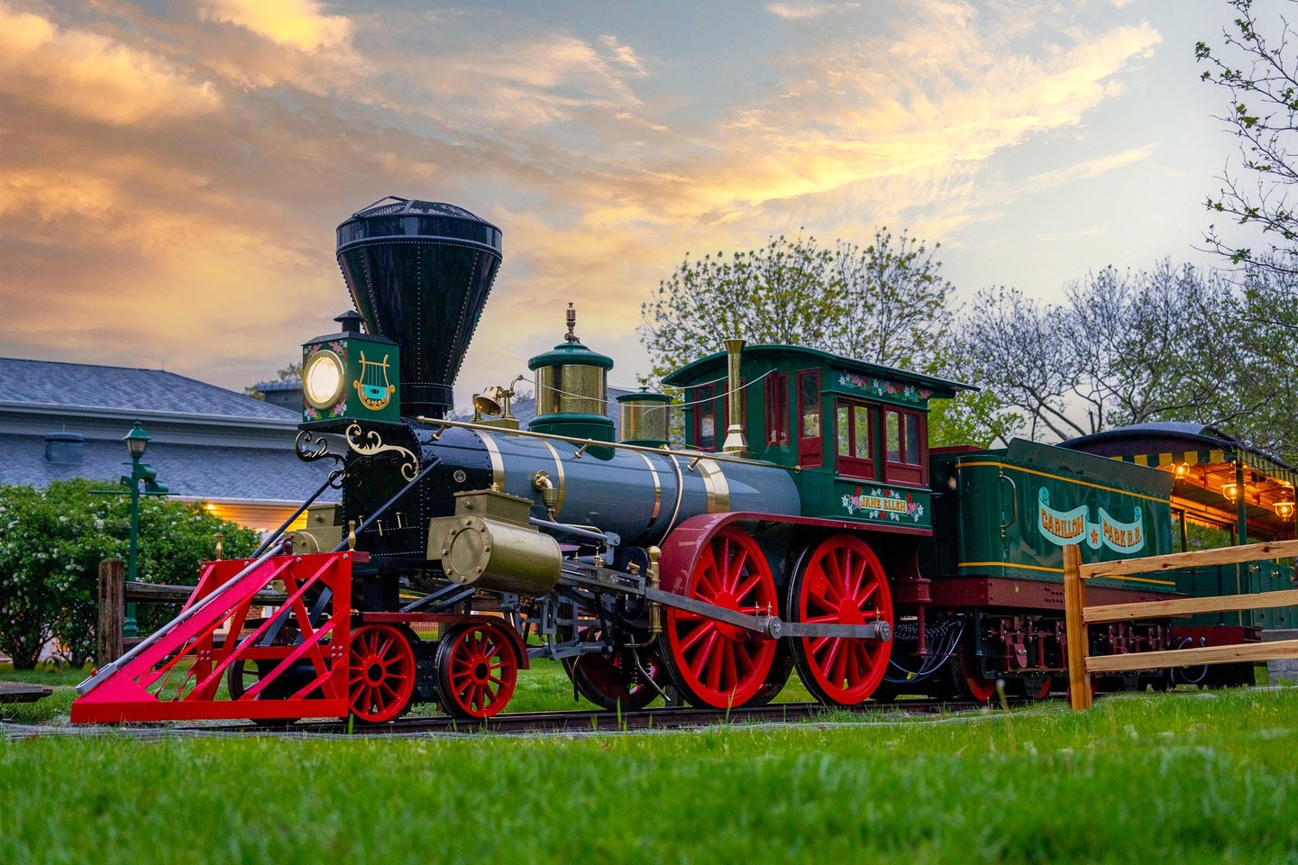 A vintage steam locomotive with red wheels and a green and black exterior sits on a grassy area, sur…