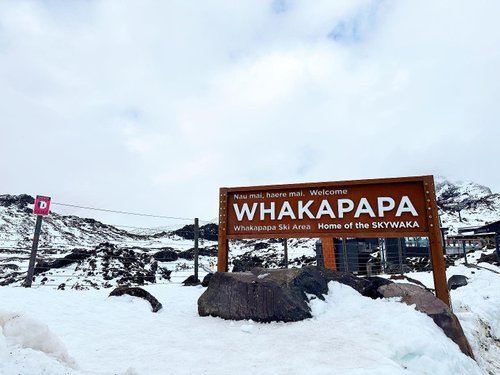Sign welcoming visitors to Whakapapa Ski Area, surrounded by snow-covered mountains and rocky terrain under a cloudy sky.