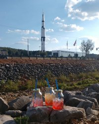 Three colorful drinks in mason jars sit on rocks in the foreground, with a tall rocket monument and flags in the background under a clear sky.