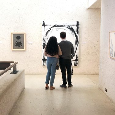 A couple stands in an art gallery, observing a large abstract painting. The minimalist space features light-colored walls and framed artworks.