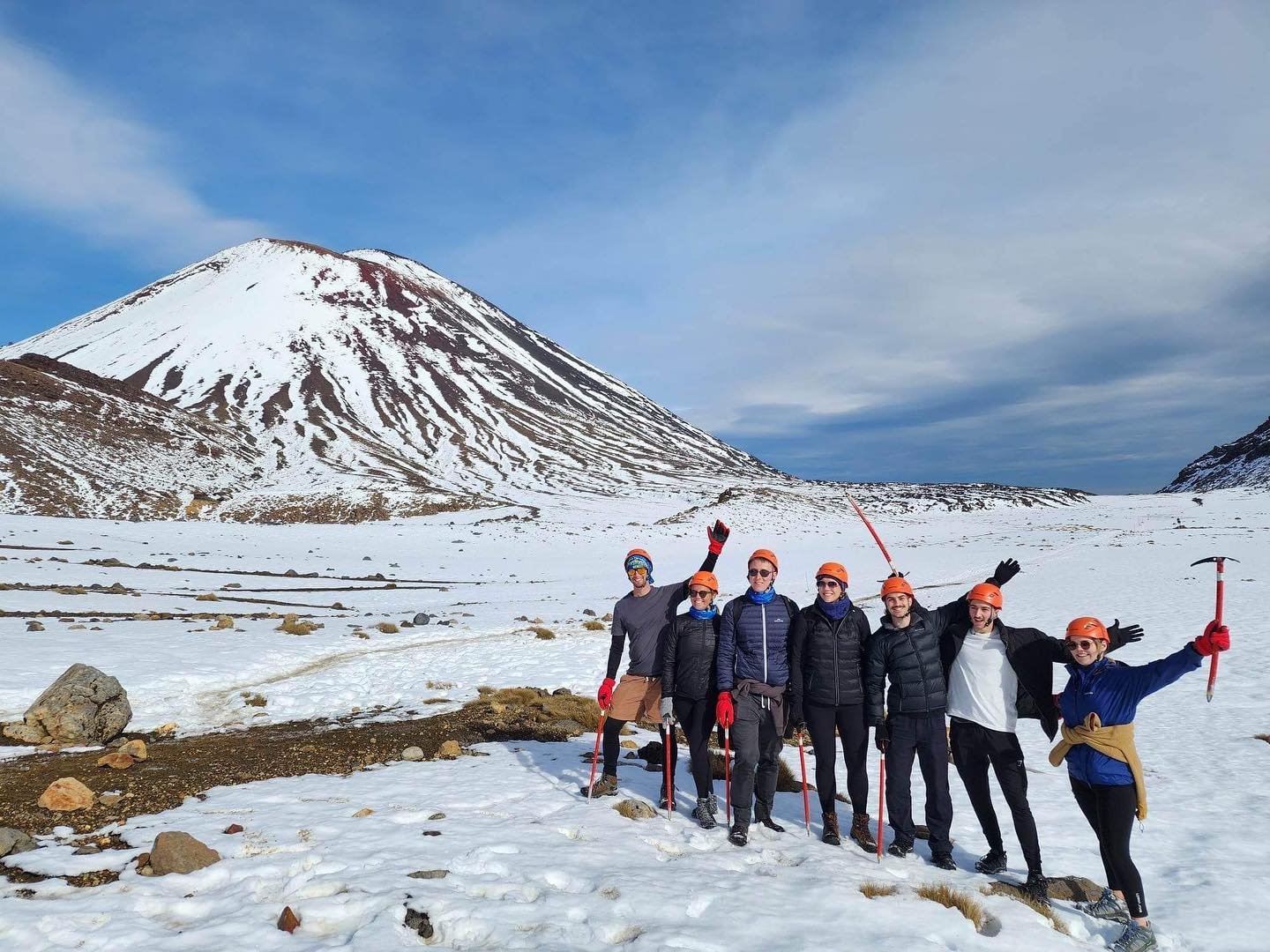 A group of seven people in winter gear stands on snow-covered terrain, with a snow-capped mountain in the background under a blue sky.
